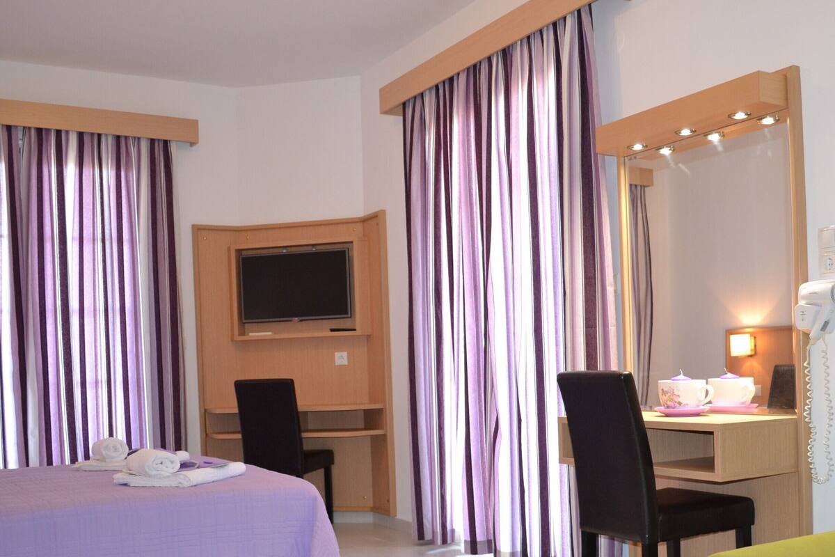 A hotel room with a bed, a desk with a flat-screen TV, a chair, a vanity with a mirror and lights, and a hairdryer.