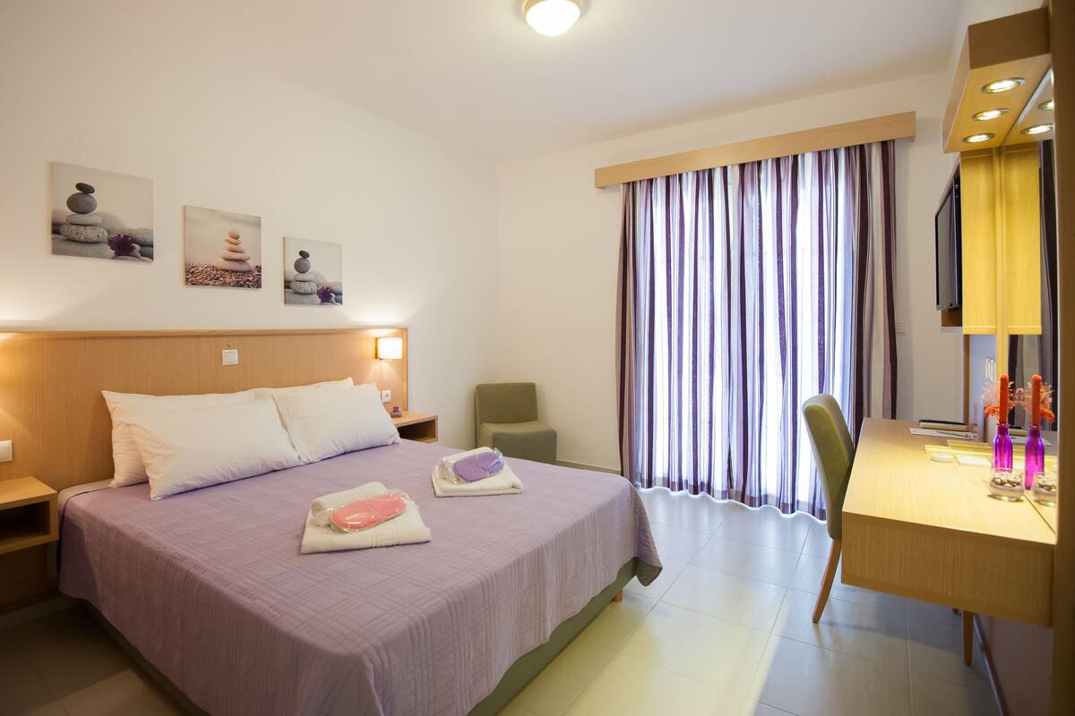 A hotel room with a bed, a desk, and a chair. There are paintings on the wall and a window with curtains.