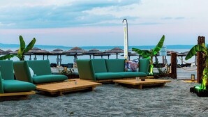 A beachside seating area with two green cushioned sofas and wooden tables, overlooking the sea with thatched umbrellas in the background. - Royal Hotel and Suites (Kassandra)