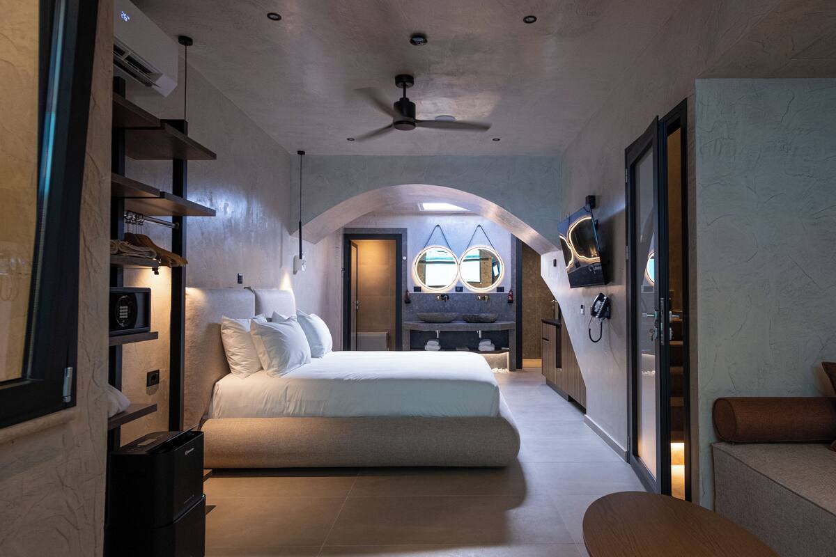 A modern hotel room with a bed, a sofa, a desk, and a bathroom with a mirror and sink.