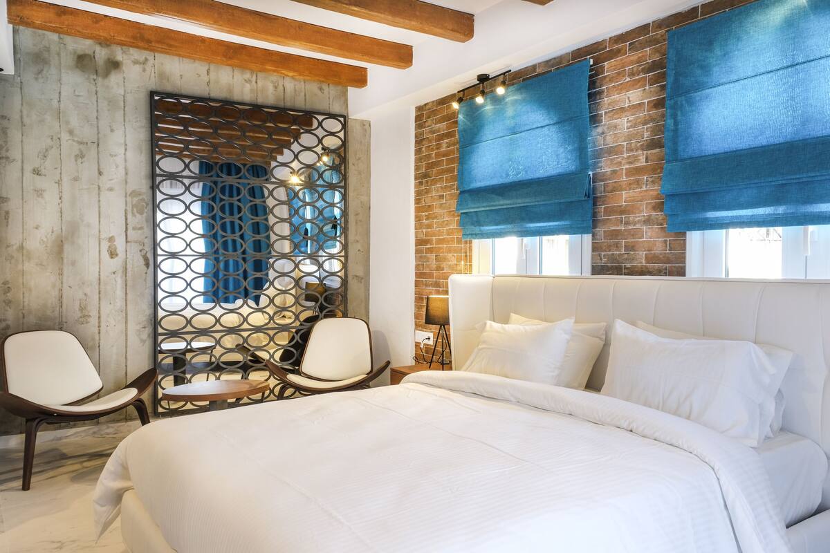 A bedroom with a large bed, two chairs, a wooden headboard, and a brick wall.