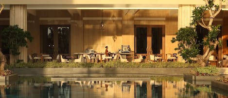A poolside area with a seating arrangement, a dining area, and a reflection in the water.