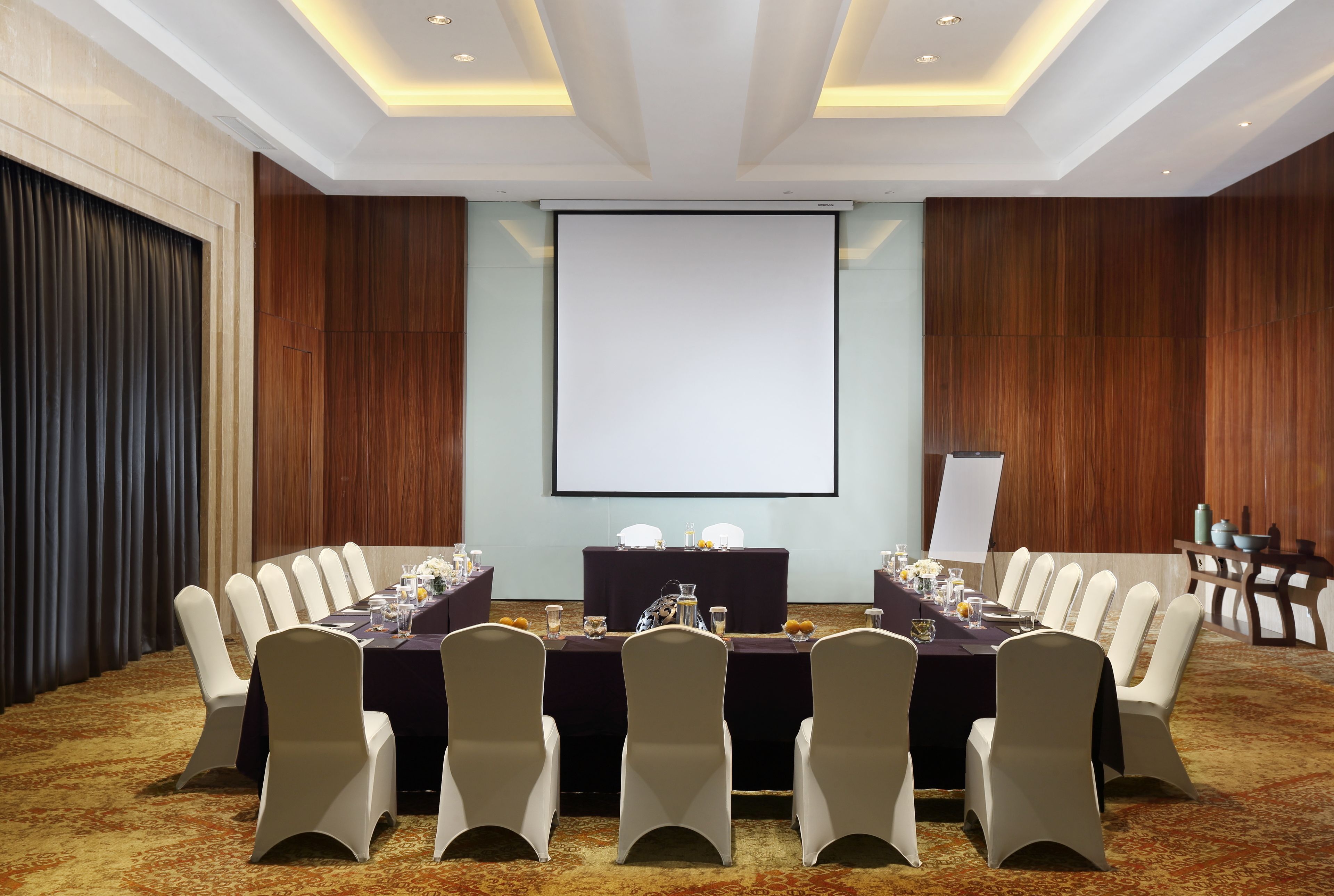 Un salón de conferencias con una gran mesa, sillas, una pantalla de proyector y una pizarra.