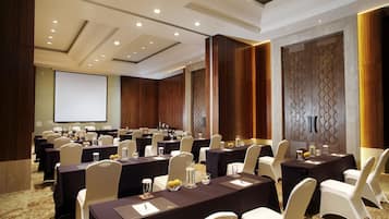 Un salón de conferencias con varias mesas y sillas, equipado con botellas de agua y blocs de notas, y una pantalla de proyector en la pared.