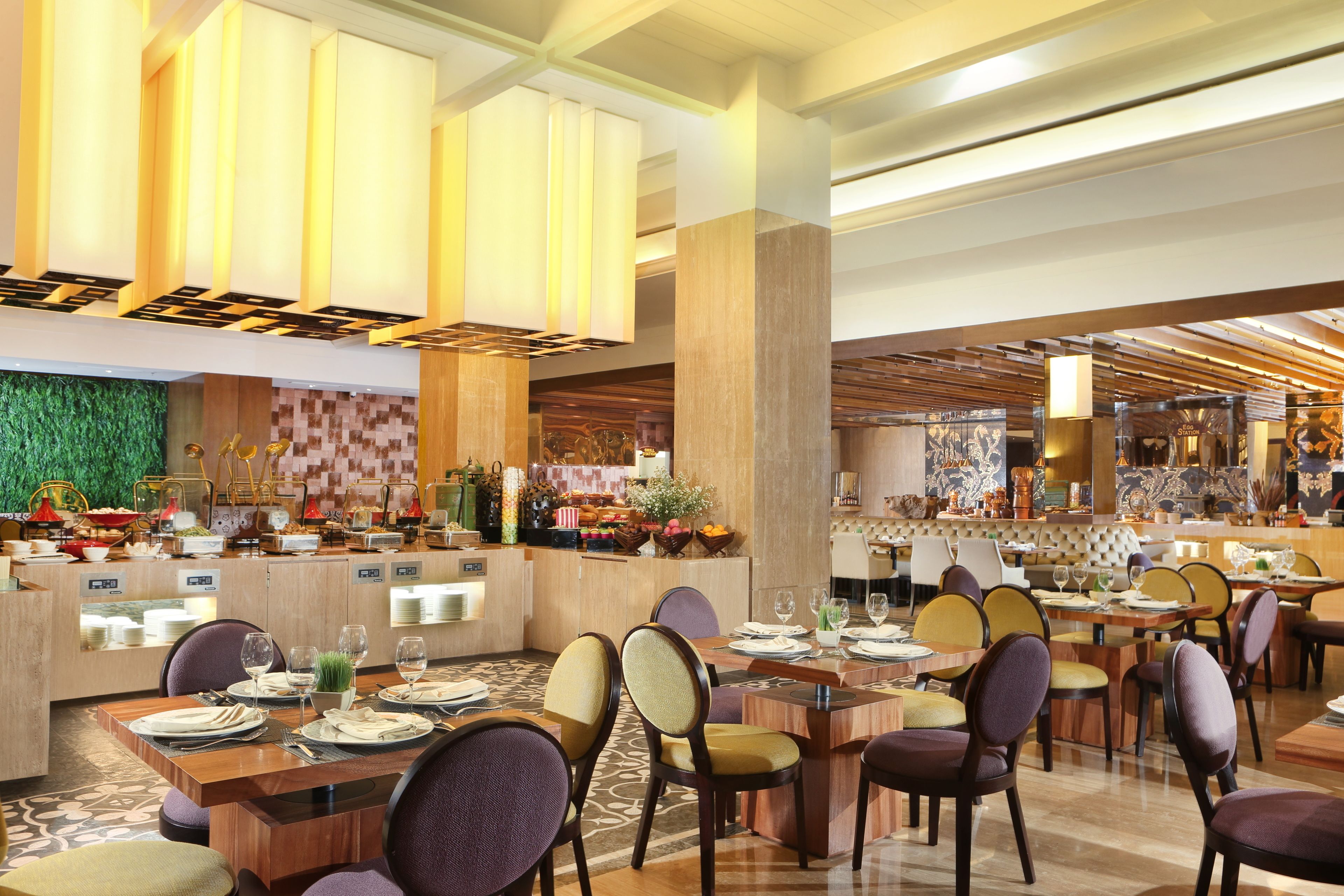 A spacious restaurant with multiple dining tables and chairs, a central bar area, and a green wall with a textured pattern.