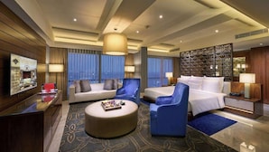 Une chambre d’hôtel moderne avec un grand lit, un canapé, un fauteuil bleu, une table basse ronde et un téléviseur placé sur le mur de gauche.