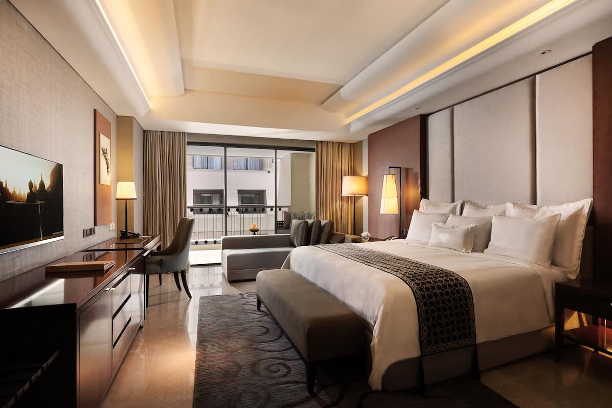Habitación de hotel con una cama grande, un escritorio, una silla, un sofá, un televisor y un balcón con vistas a edificios.