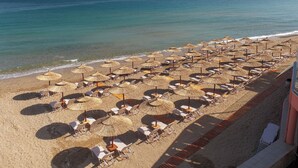 A beach with rows of sun loungers and thatched umbrellas, a clear view of the ocean, and a sandy walkway along the beach. - Dedalos Beach Hotel (Rethymno)