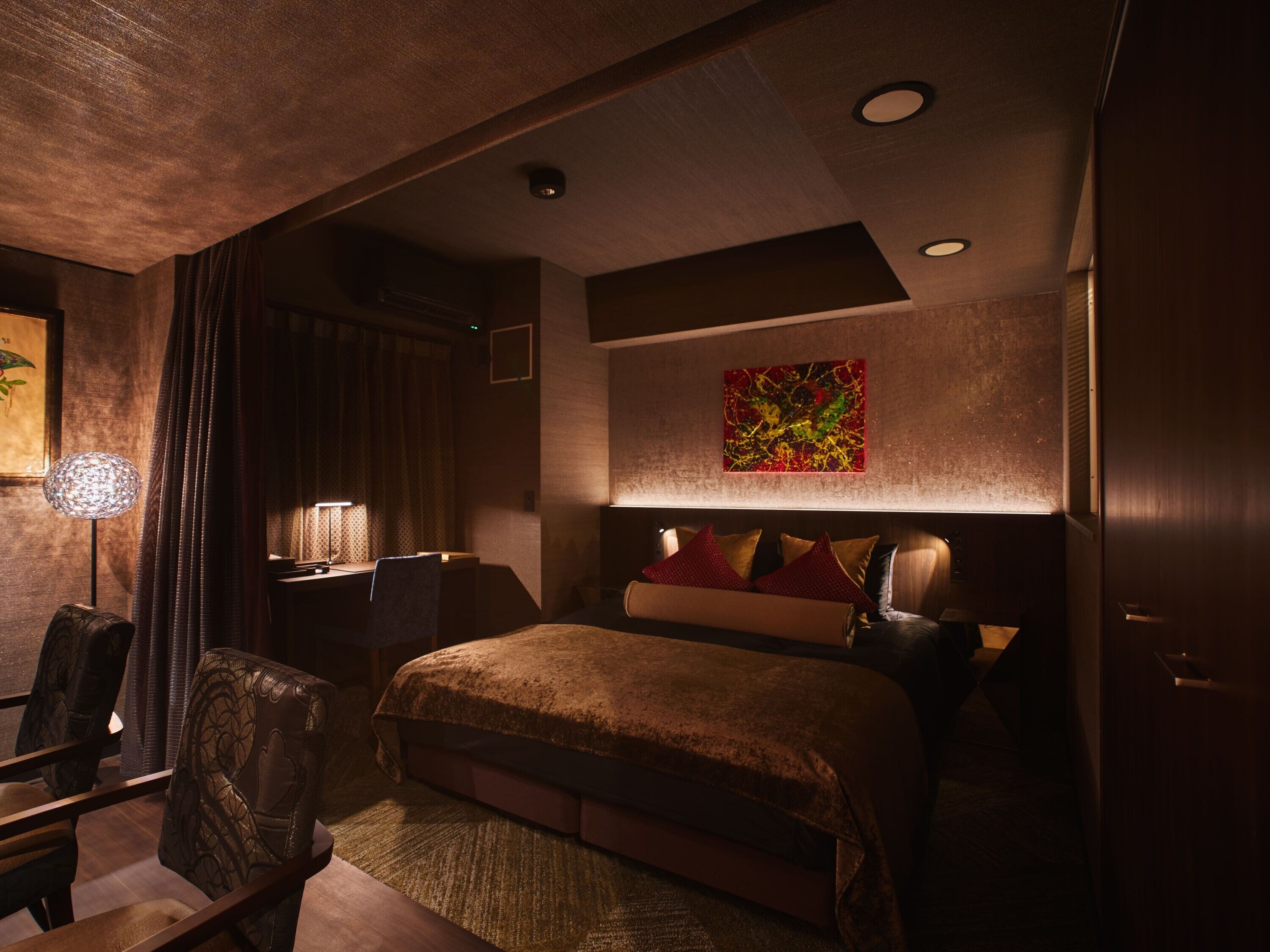 A dimly lit hotel room with a large bed, a desk with a lamp, and a painting on the wall.