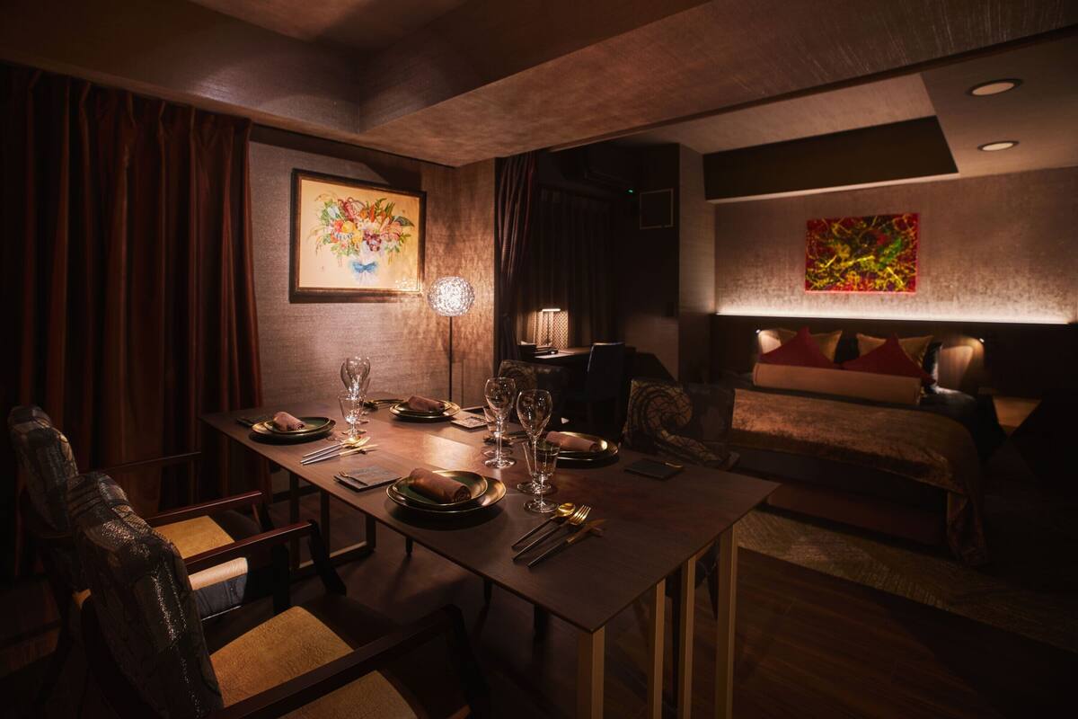 Een gedempt verlichte eetruimte met een lange tafel gedekt voor vier personen, omringd door stoelen. Een schilderij hangt aan de muur, en een lamp zorgt voor licht.