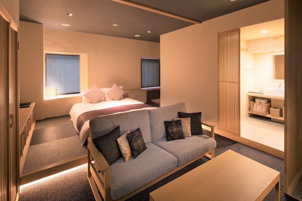 Een moderne hotelkamer met een bank, een bed, een kleine tafel en een badkamer die door een open deur zichtbaar is.