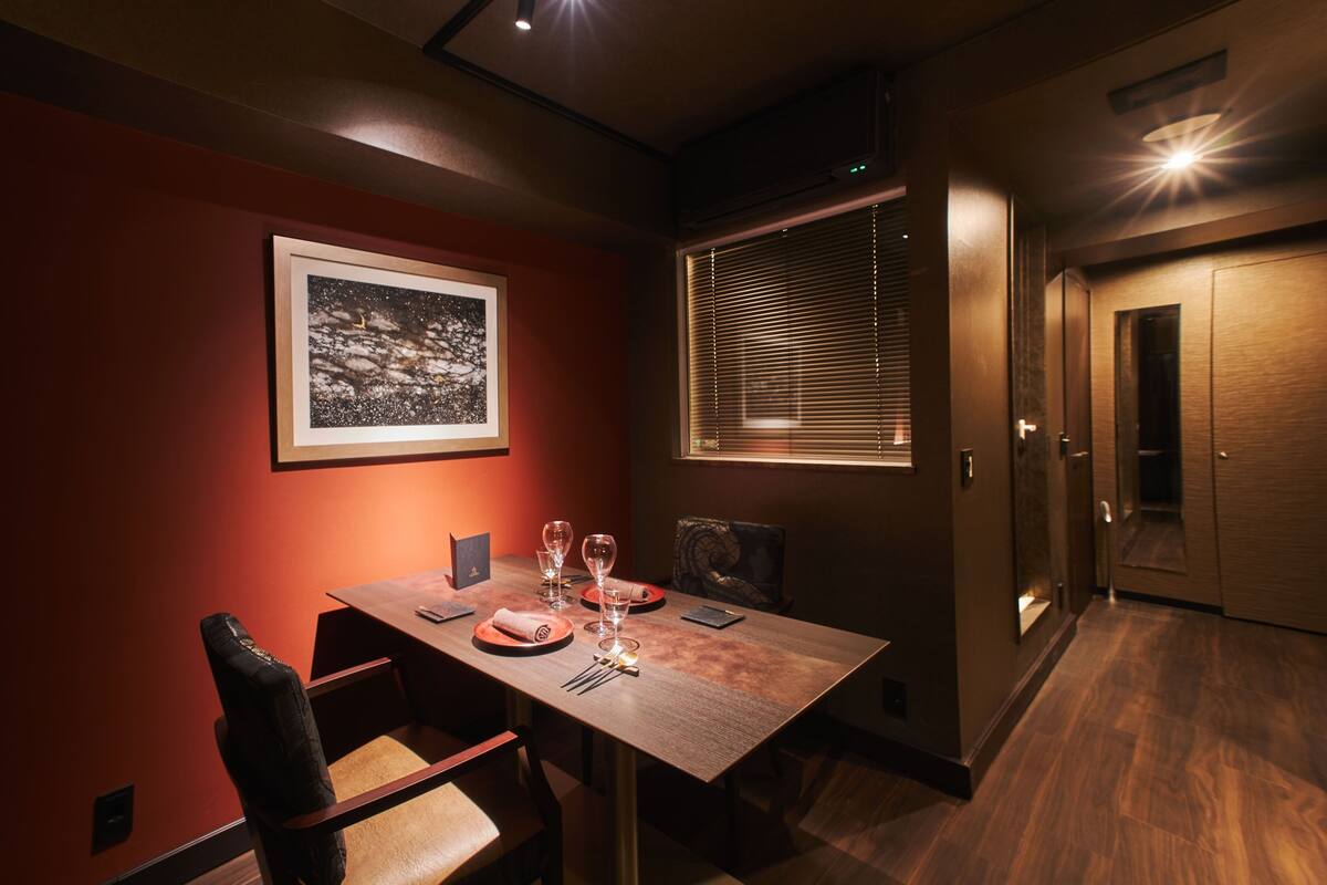 Een eetgedeelte met een tafel gedekt voor twee personen, een lijst met een afbeelding aan de muur en een raam met zonwering.