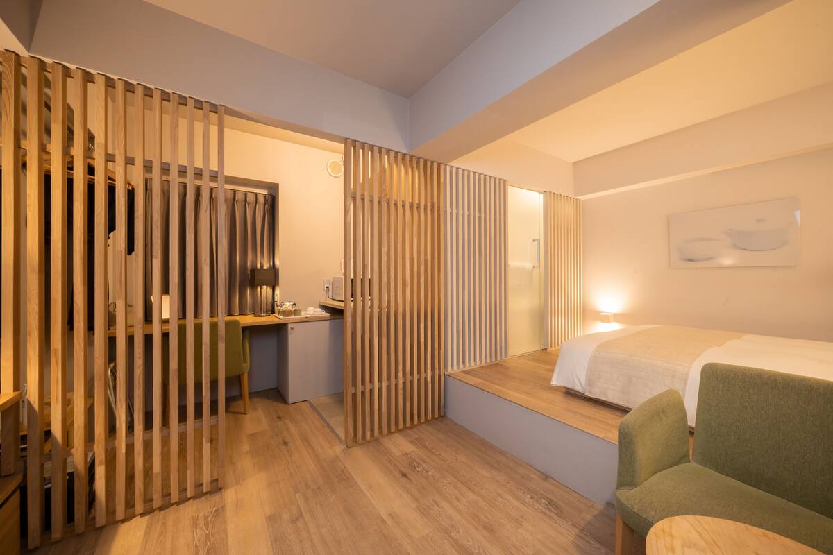 Een moderne hotelkamer met een houten scheidingswand, een bed, een groene fauteuil en een bureau met een lamp.