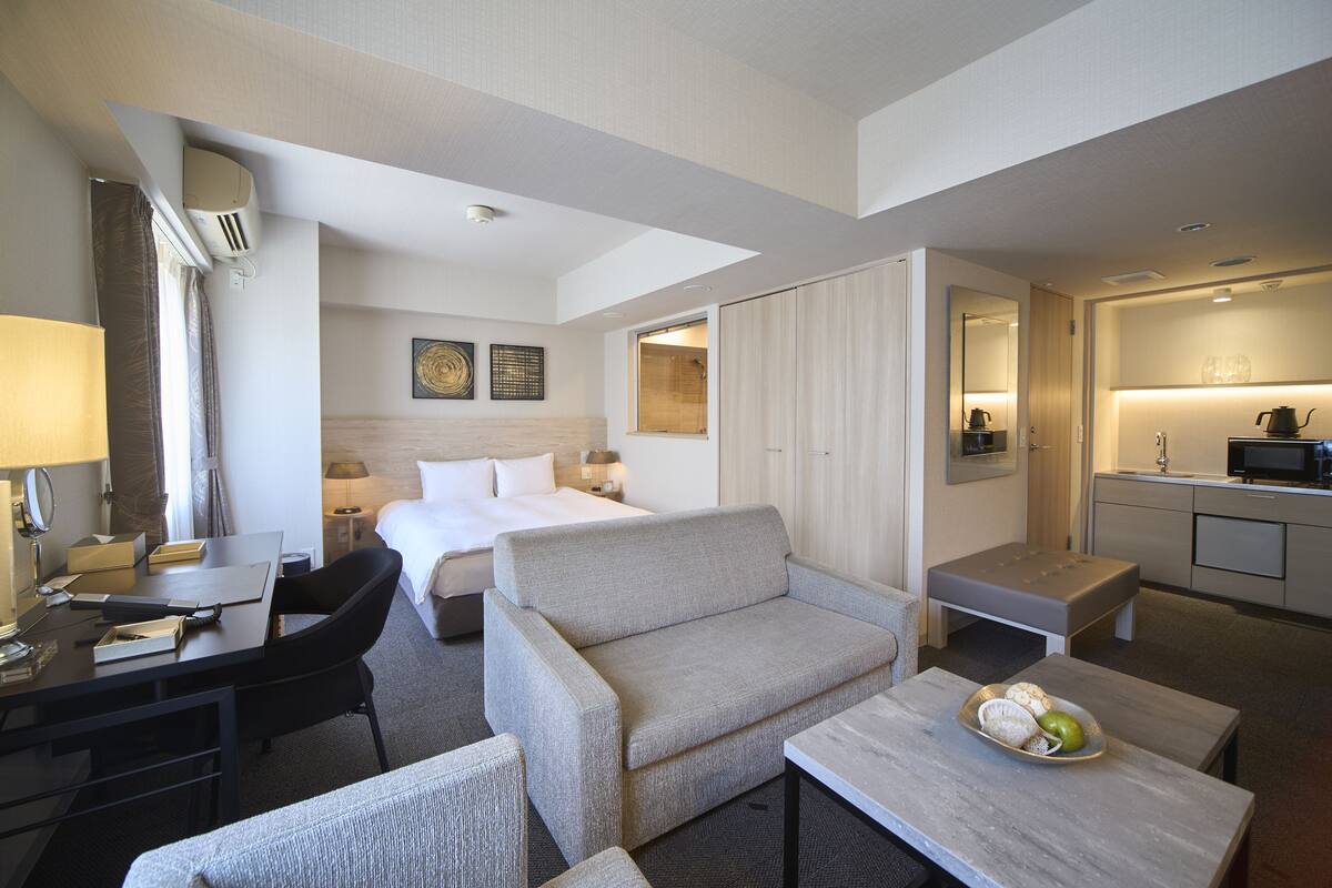 Een hotelkamer met een bed, een bank, een bureau, een kleine tafel met fruit, een kitchenette en een badkamer.