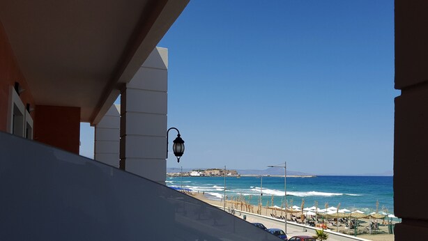 A balcony with a view of the sea, a street with parked cars, and a distant shoreline with buildings. - Blue Sky Apartments (Rethymno)