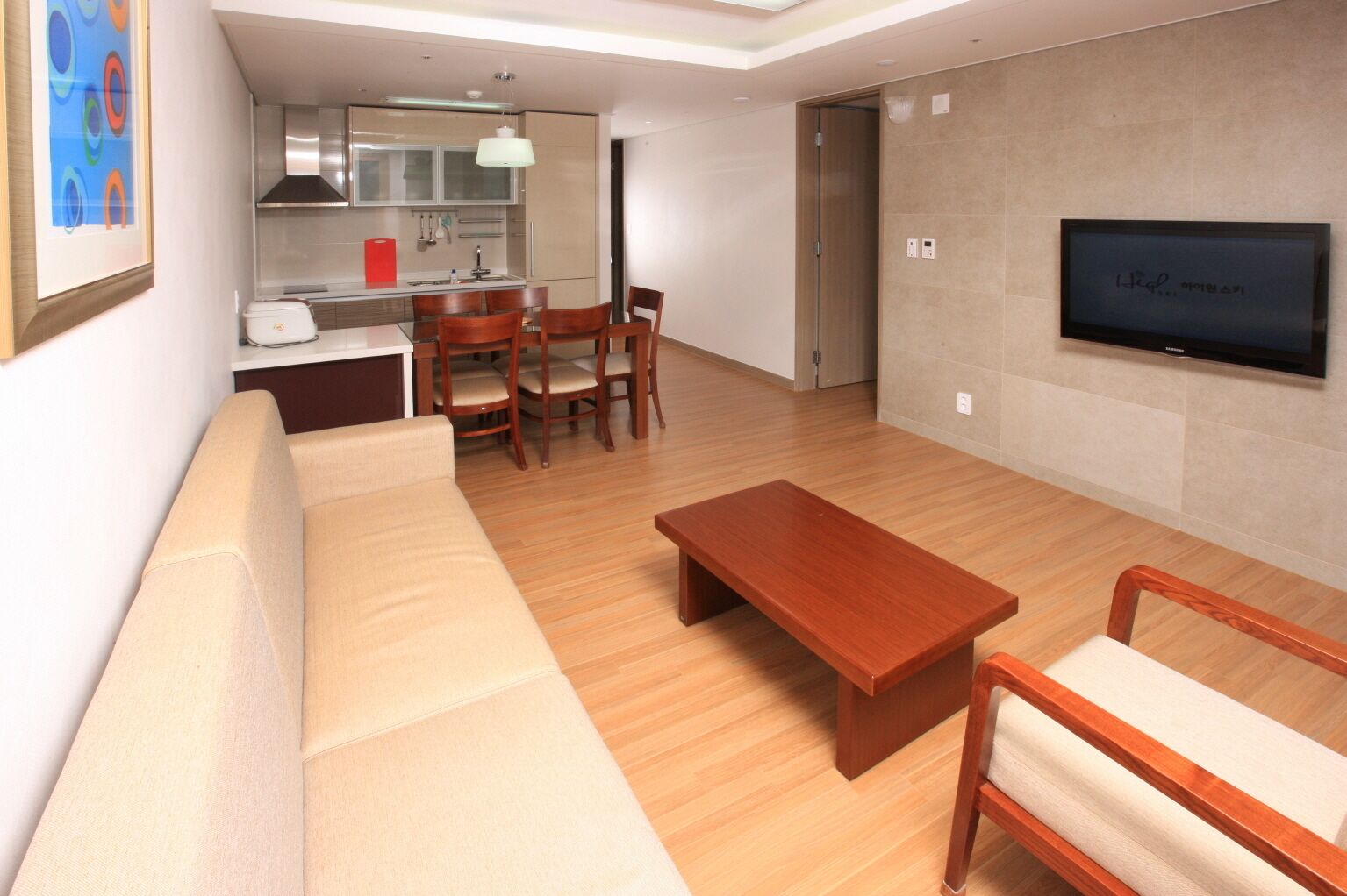 Uma sala de estar moderna com sofá, mesa de centro de madeira, mesa de jantar com cadeiras, área de cozinha e TV fixada na parede.