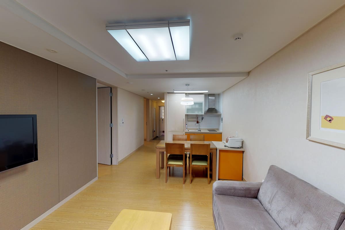 Ein modernes Hotelzimmer mit Flachbildfernseher, einer Couch, einem Esstisch mit Stühlen, einer Küchenzeile und einem Flur.
