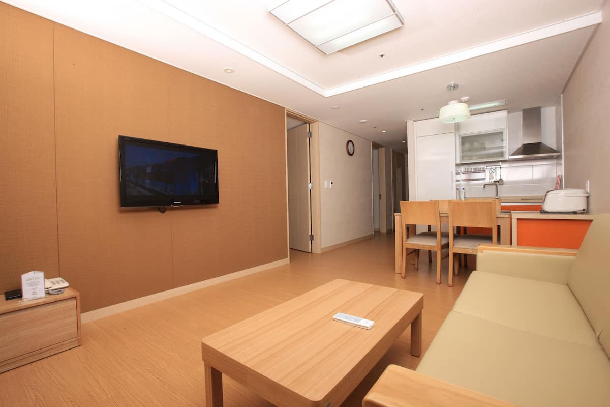 Ein modernes Hotelzimmer mit Holzboden, einem Wand-Flachbildfernseher, einem Holztisch, einem beigen Sofa und einer Küchenzeile mit Spüle und Herd.