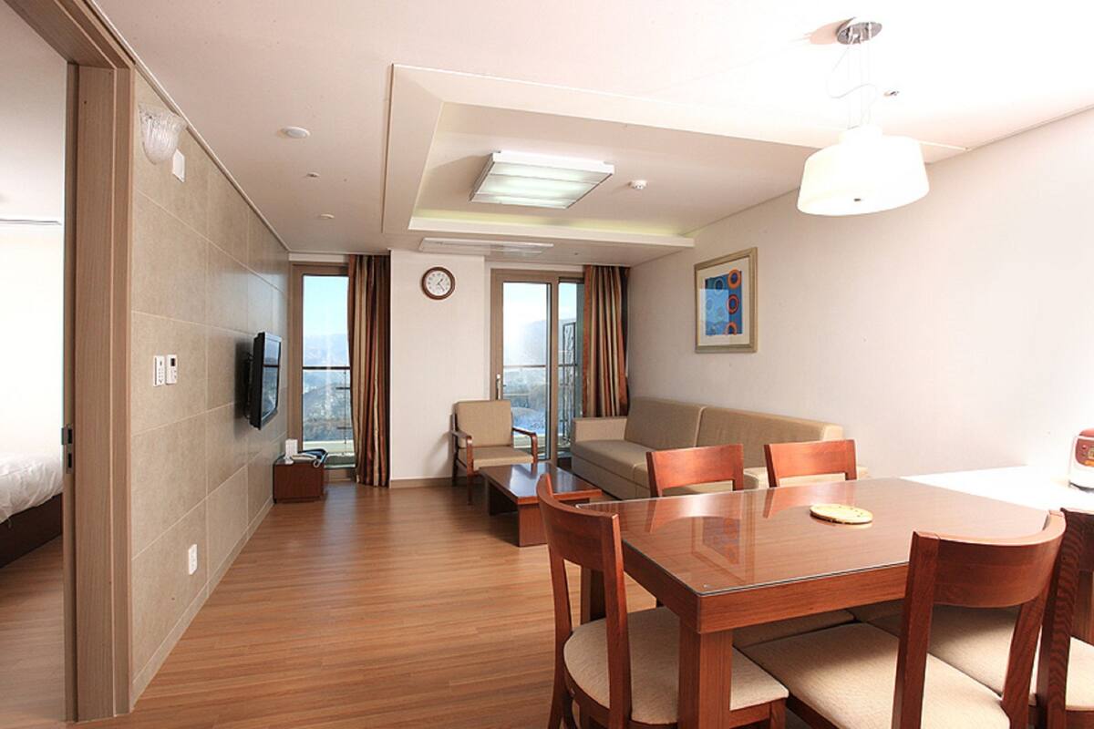 Ein Hotelzimmer mit Bett, Fernseher, Esstisch mit Stühlen, einer Couch und einem großen Fenster mit Vorhängen.