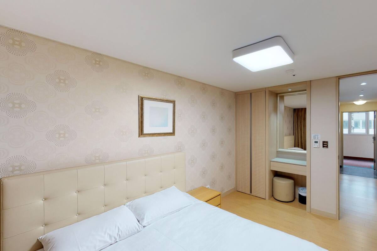 Ein Hotelzimmer mit einem Bett, einem Nachttisch, einem Spiegel, einer Lampe und einer Tür zu einem anderen Raum.