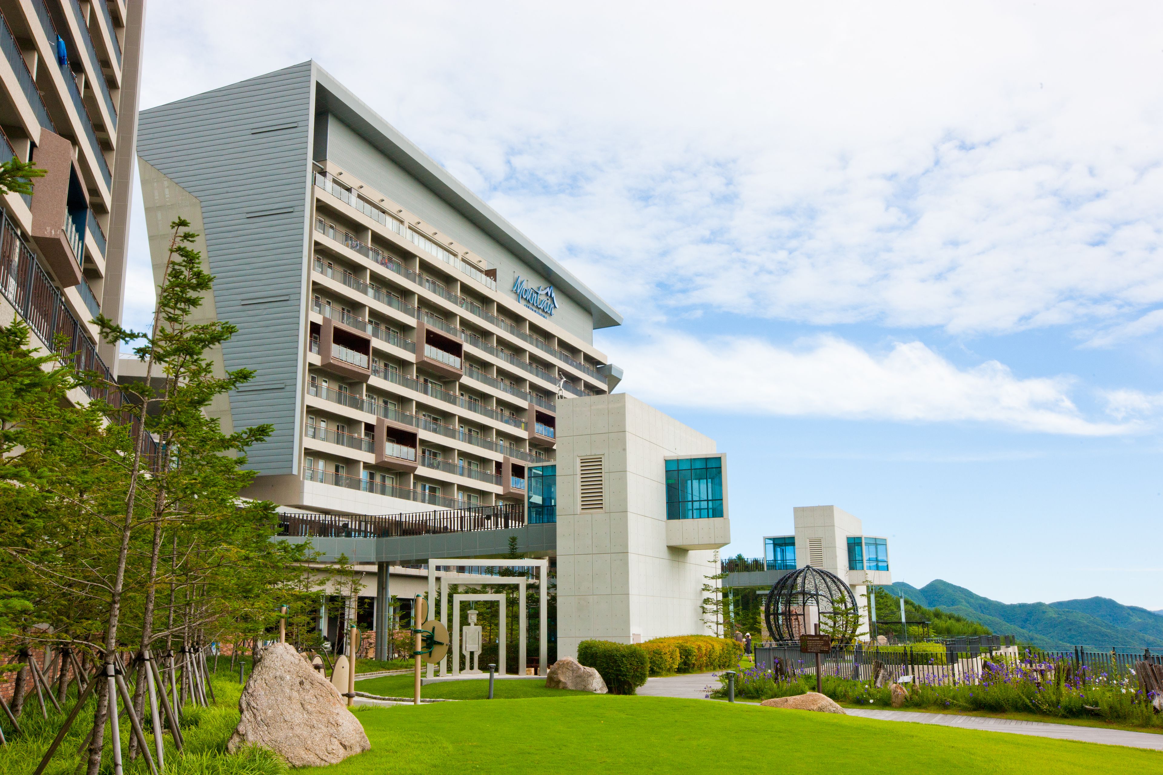 A modern hotel with multiple floors, a well-maintained lawn, and a view of mountains in the background.