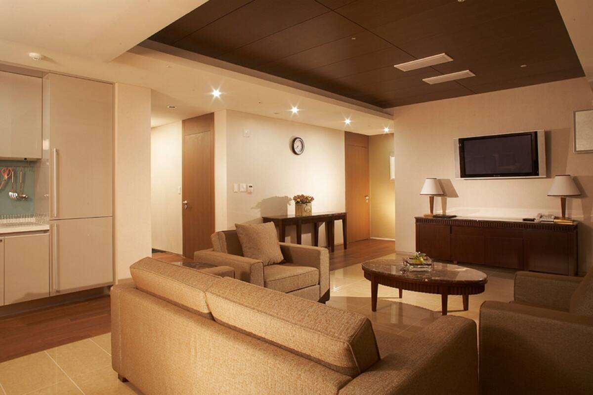 Ein modernes Wohnzimmer mit einer Couch, Sesseln, einem Couchtisch und einem Fernseher. An der Wand hängt eine Uhr und ein Schrank mit Küchenutensilien steht dort.