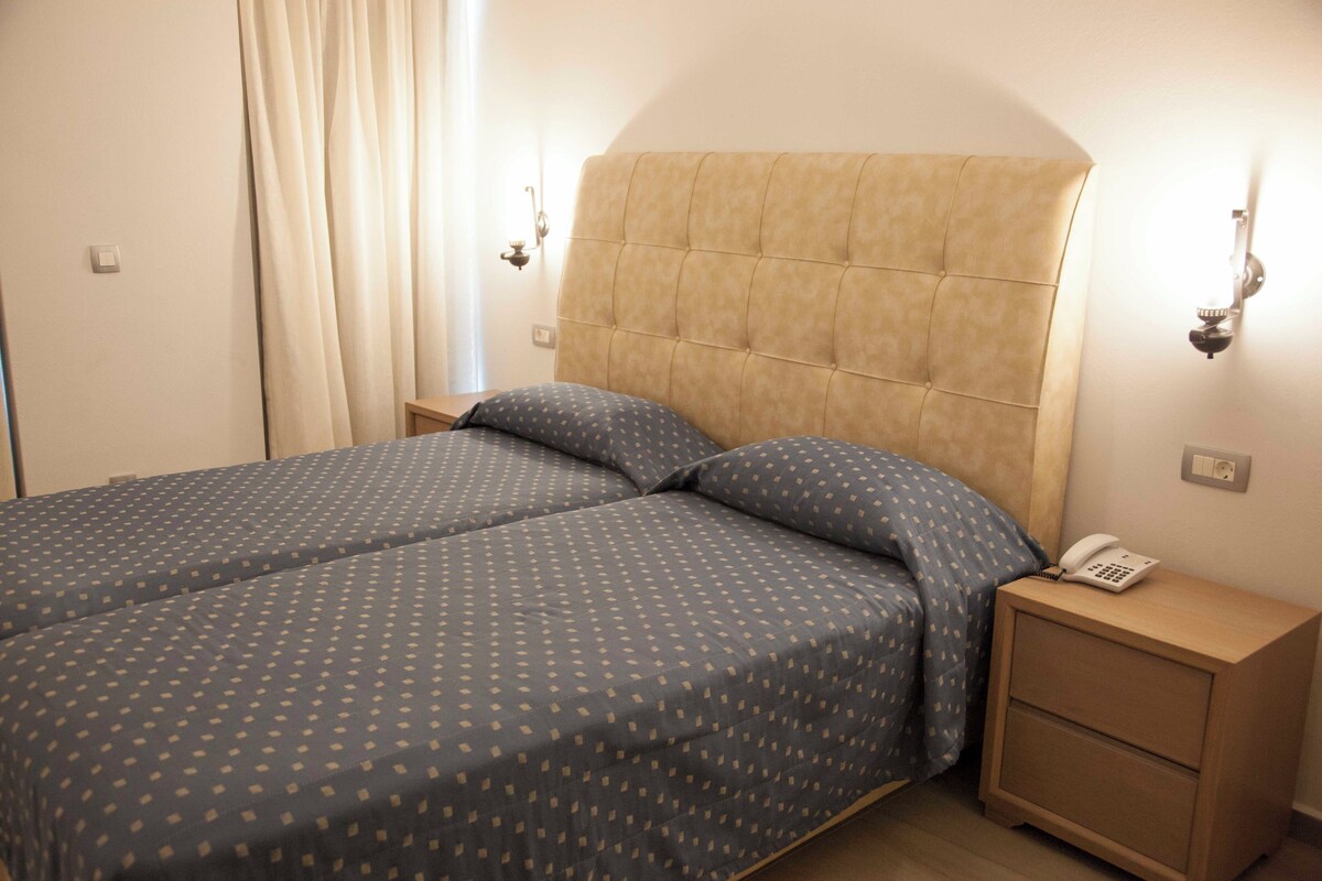 A double bed with a tufted headboard, a bedside table with a telephone, and a curtain on the window.