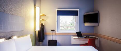 Habitación de hotel con cama, escritorio con computadora portátil, televisión, maleta y ventana con vista a follaje.