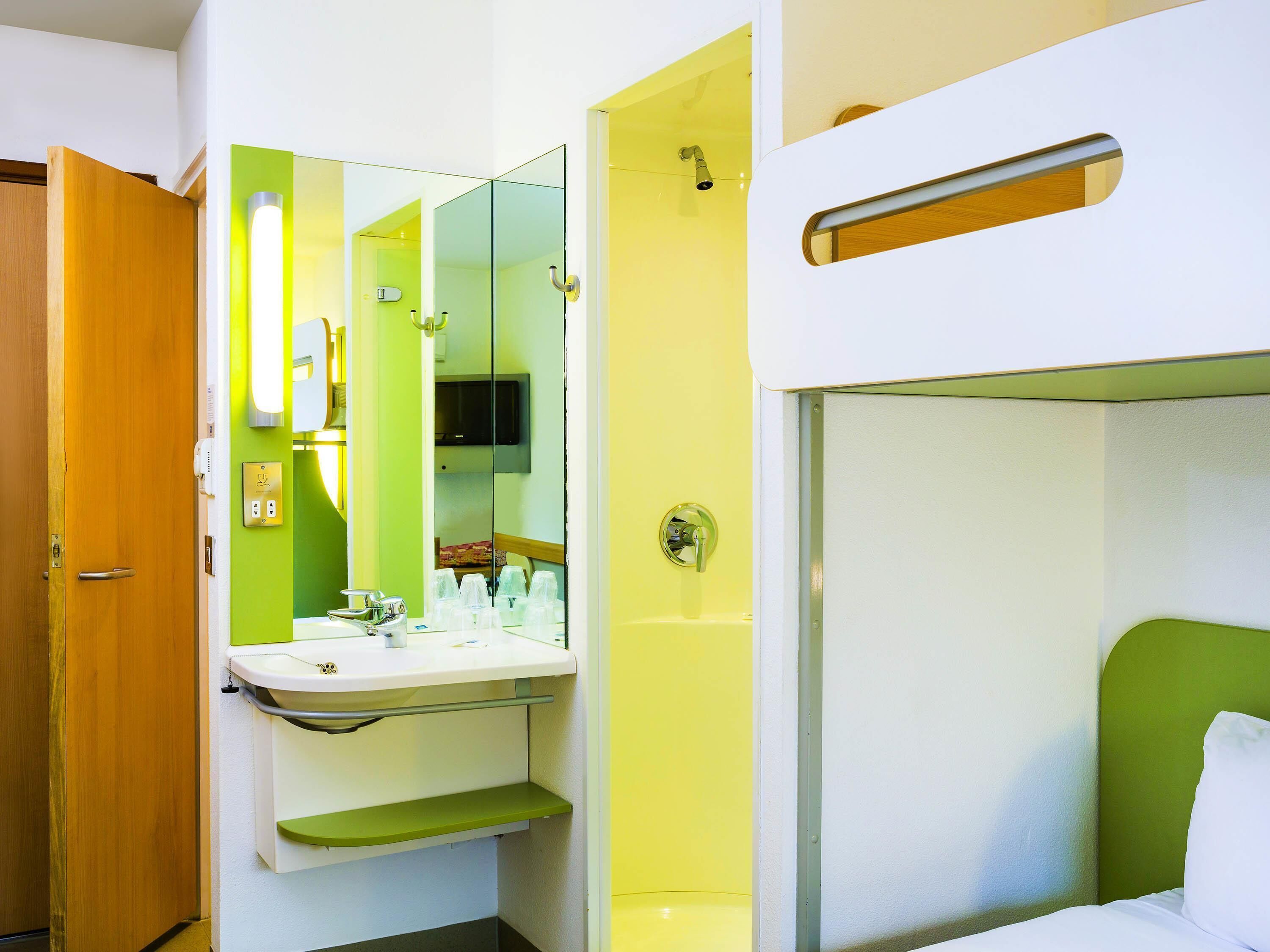 Een kleine hotelkamer met een enkel bed, een badkamer met wastafel en spiegel, en een deur die naar een andere kamer leidt.