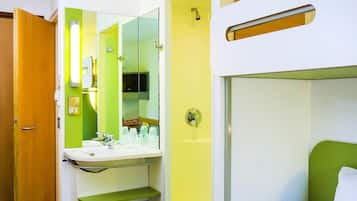 Een kleine hotelkamer met een enkel bed, een badkamer met wastafel en spiegel, en een deur die naar een andere kamer leidt.