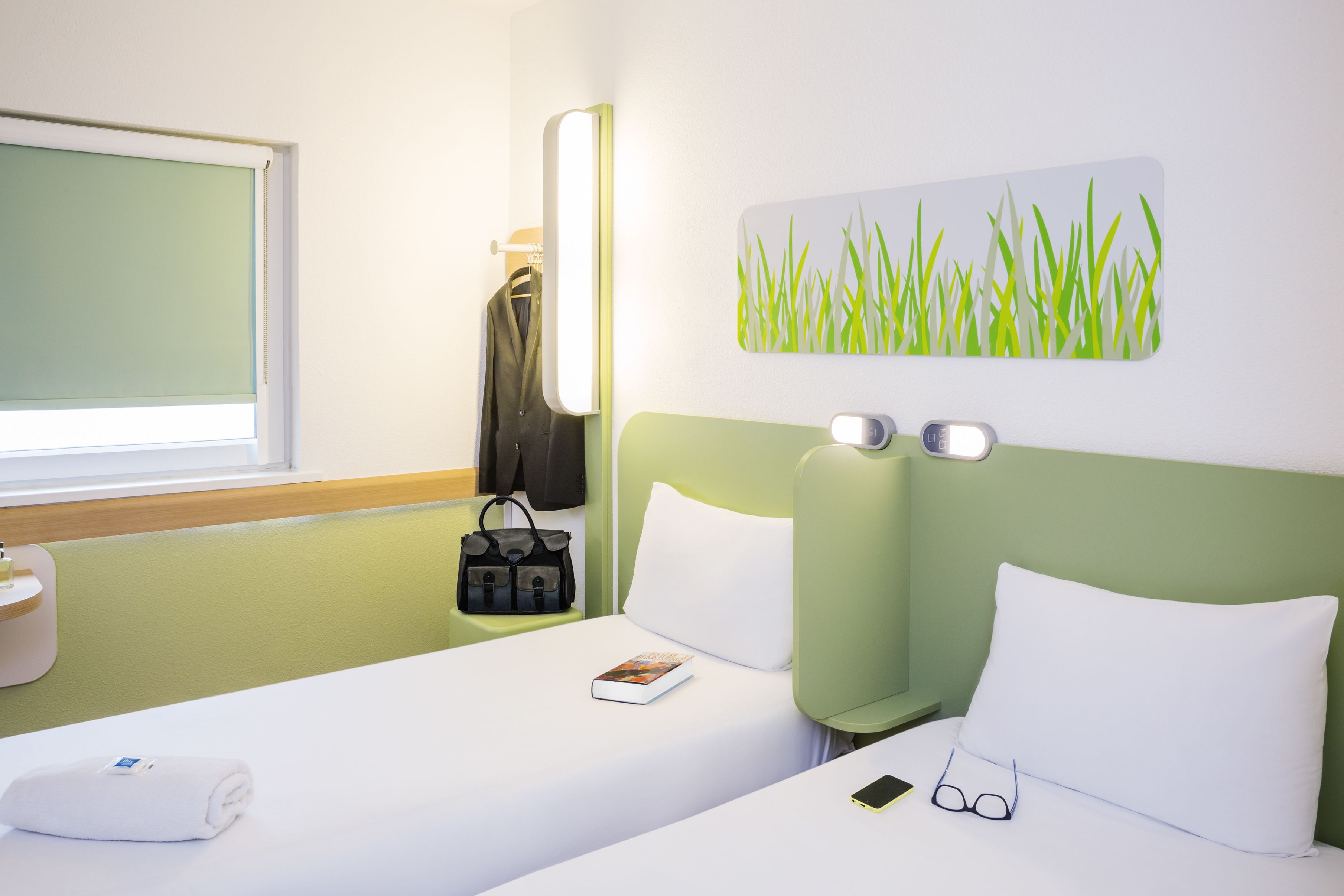 Habitación de hotel con cama, mesita de noche, lámpara de pared, un cuadro de césped y una ventana con persiana.