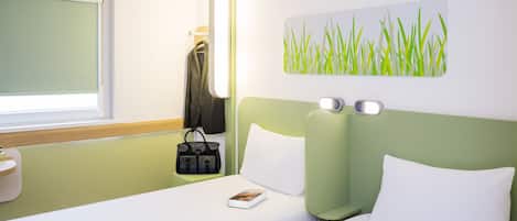 A hotel room with a bed, a bedside table, a nightstand, a wall-mounted lamp, a painting of grass, and a window with a blind.
