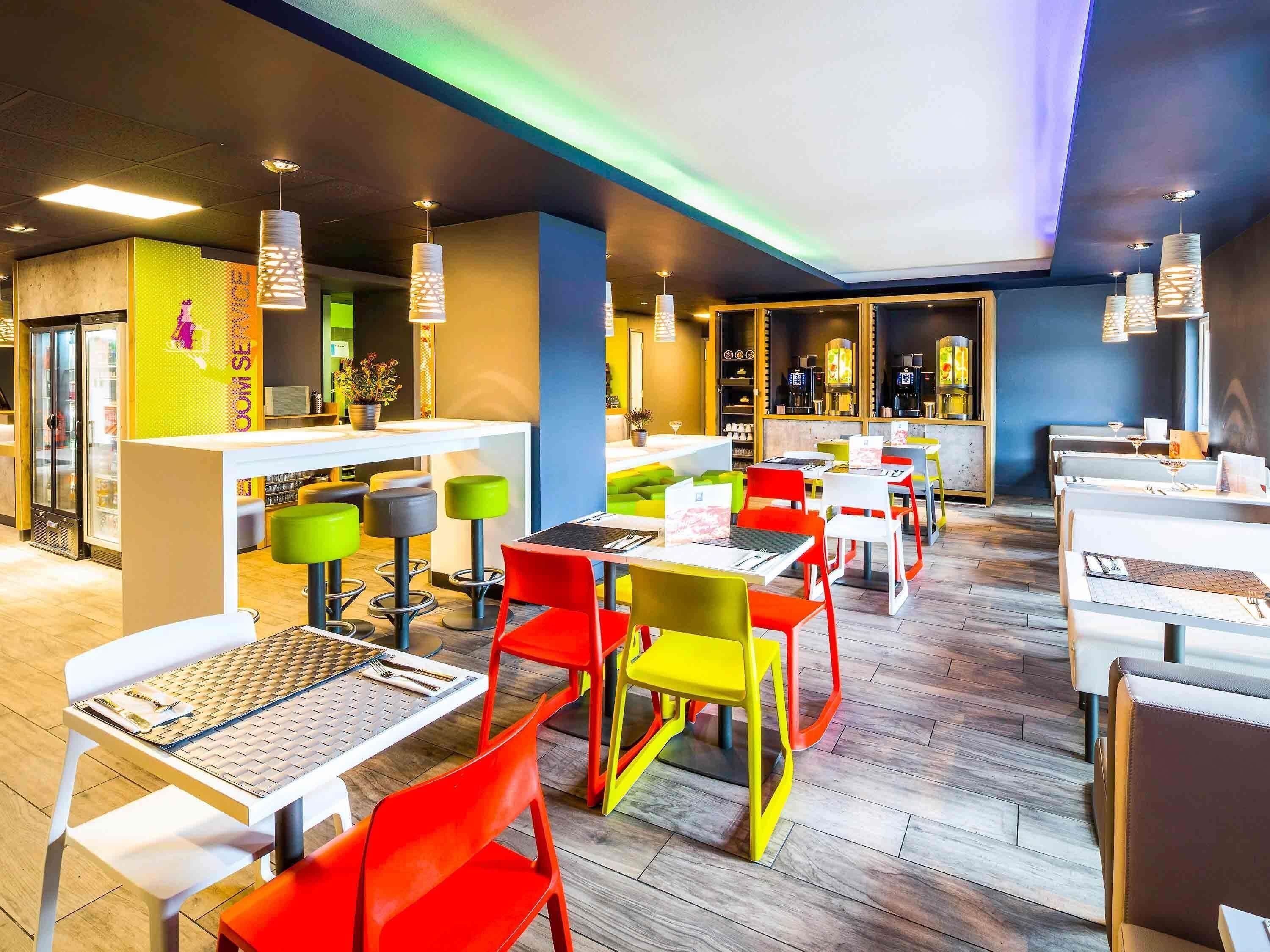 Un interior de restaurante moderno con varias mesas de comedor y sillas coloridas, una zona de bar e iluminación colgante.