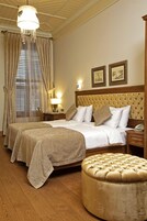 Ein Hotelzimmer mit zwei Betten, einem gepolsterten Kopfteil, einem runden Hocker, einem Nachttisch aus Holz, einer Lampe und einem Fenster mit Vorhängen.