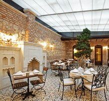Ein Restaurant mit Backsteinwänden, einer Glasdecke und verzierten Metallstühlen. Die Tische sind mit weißen Tischdecken und Servietten gedeckt.
