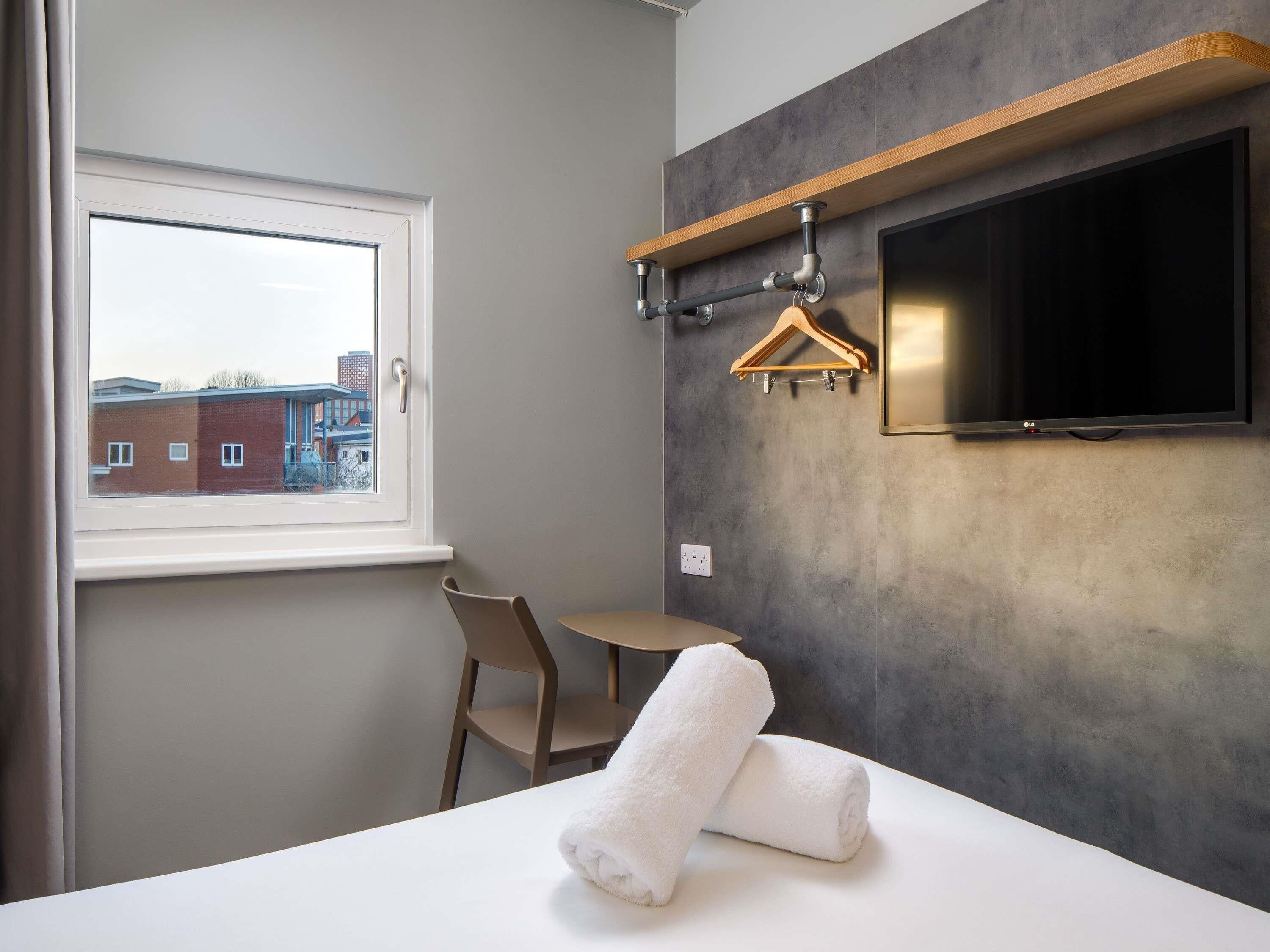 A modern hotel room with a bed, a TV mounted on the wall, a wooden hanger, and a window with a view of buildings.