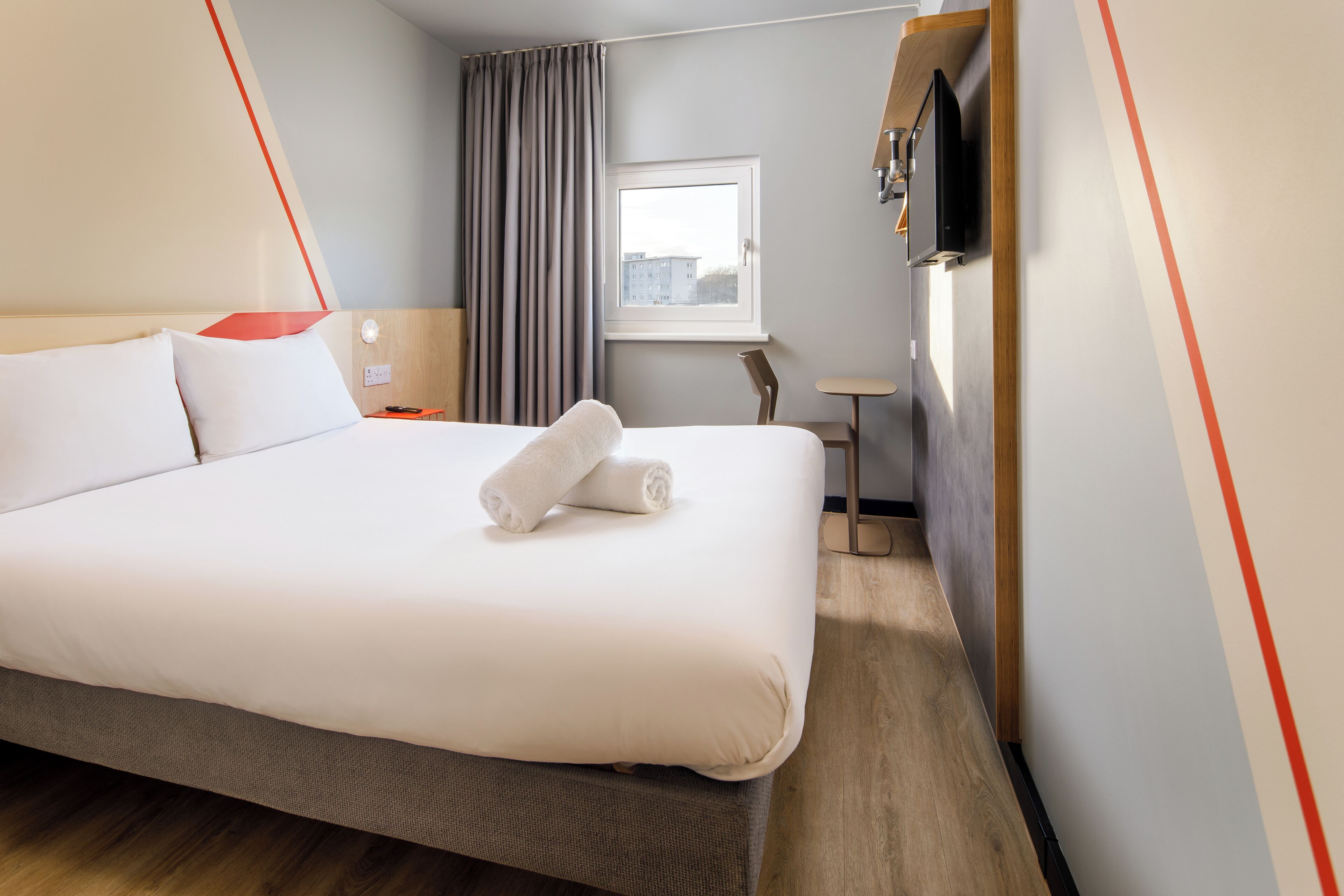 A hotel room with a large bed, a small table, a TV mounted on the wall, and a window with curtains.