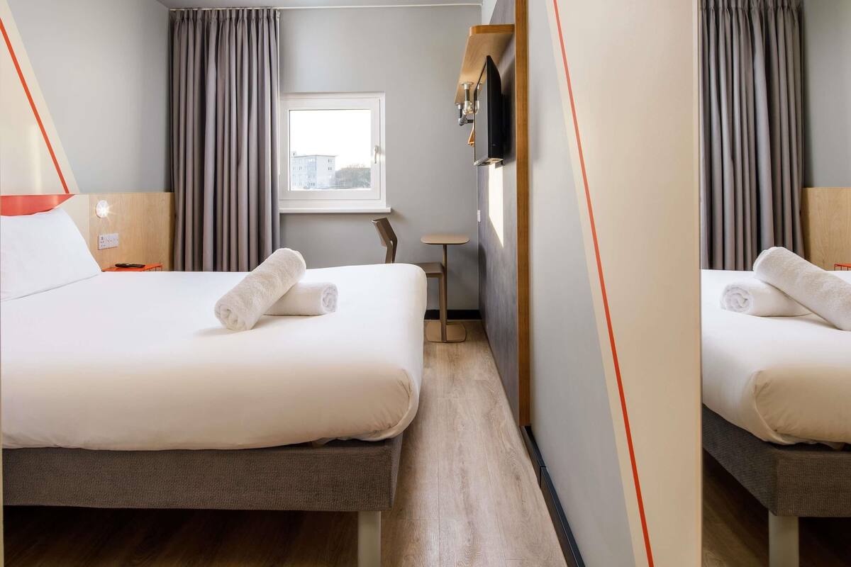 A hotel room with two beds, a small table, a TV, and a window with curtains.