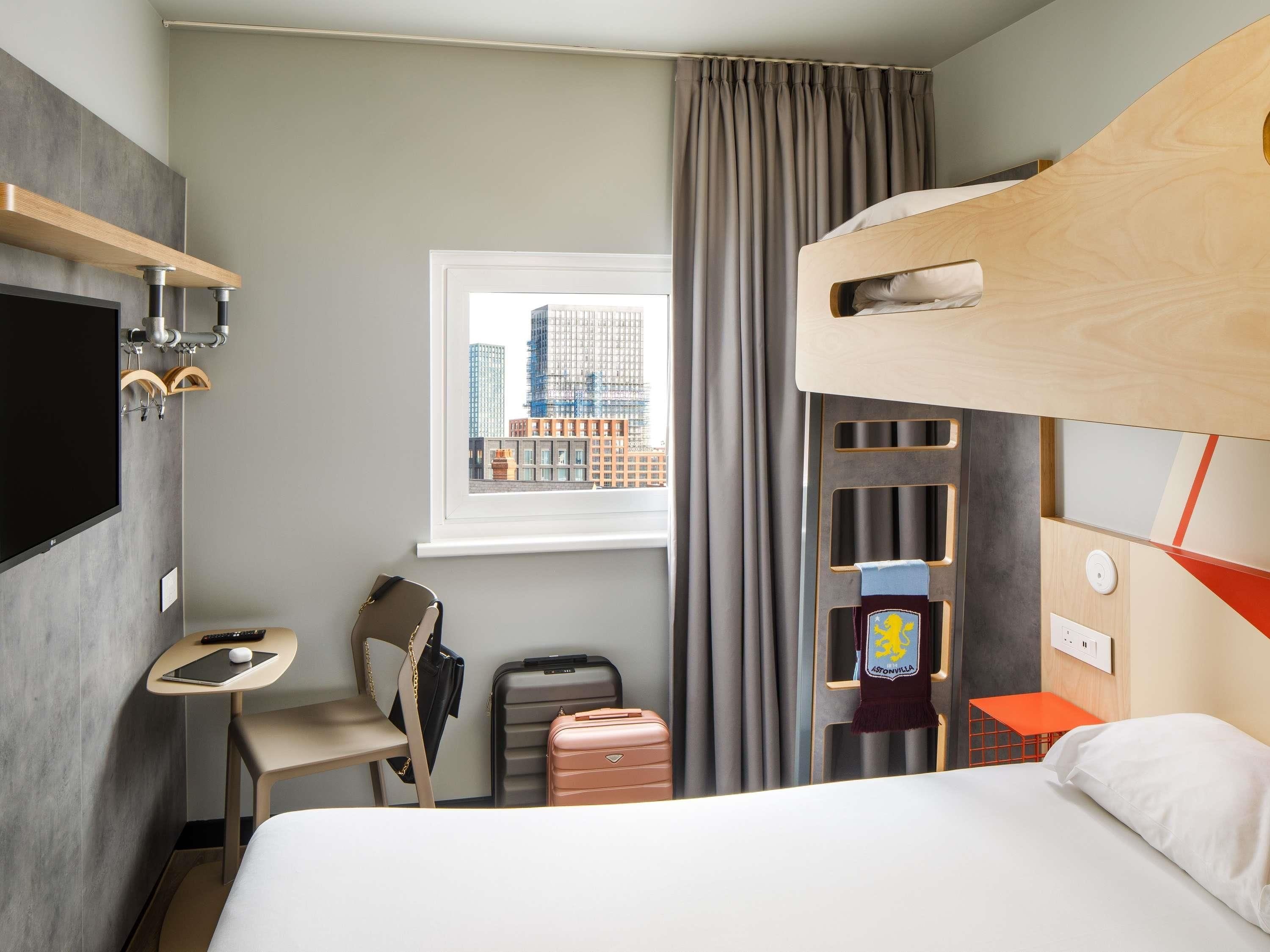 A hotel room with a bunk bed, a desk with a chair, a suitcase, a television, and a window with a city view.
