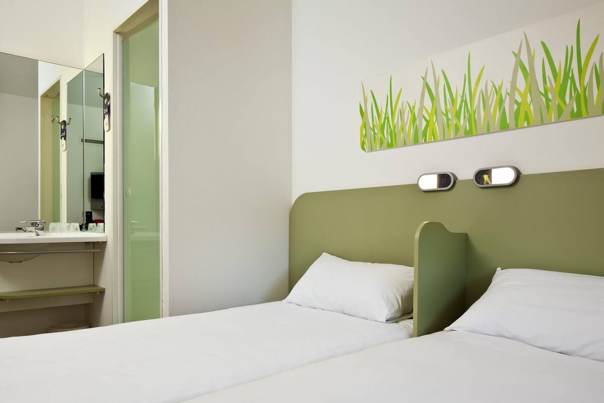 A hotel room with a bed, a bedside table, a mirror, and a wall-mounted light fixture.