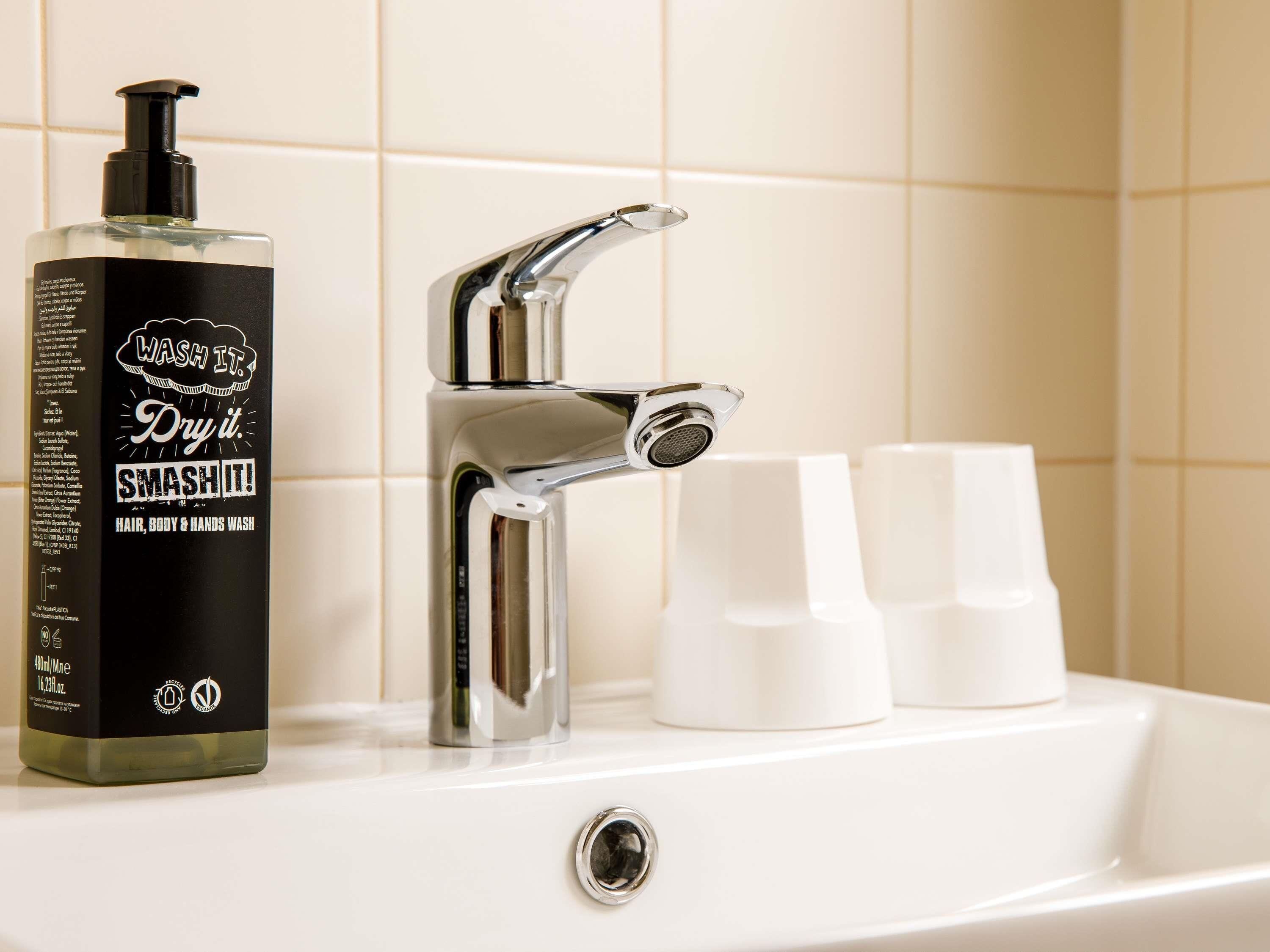 Un baño con un grifo cromado, una botella de jabón líquido y dos tazas blancas en el lavabo.