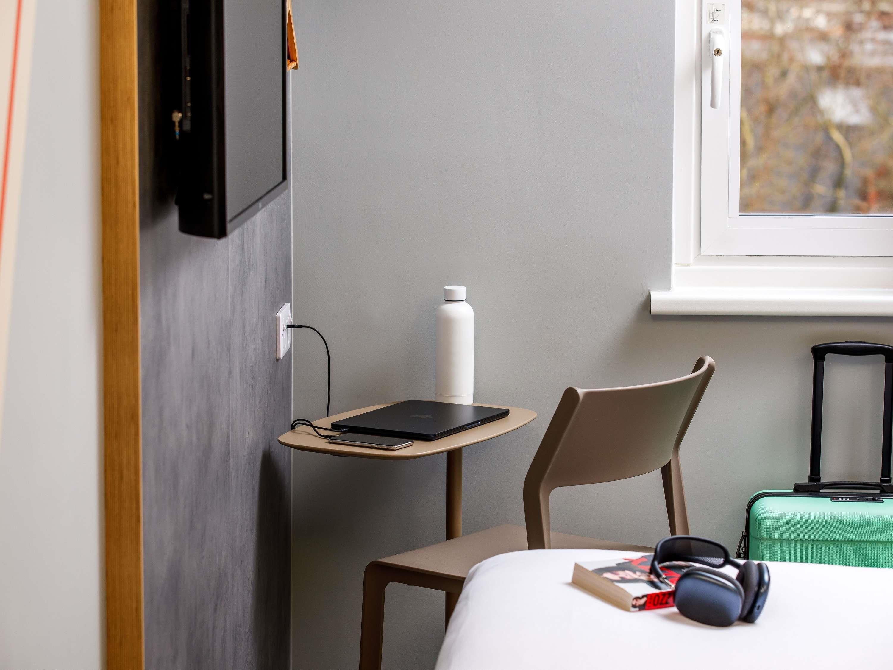 A hotel room with a bed, a chair, a small table with a laptop, a bottle, headphones, and a suitcase.