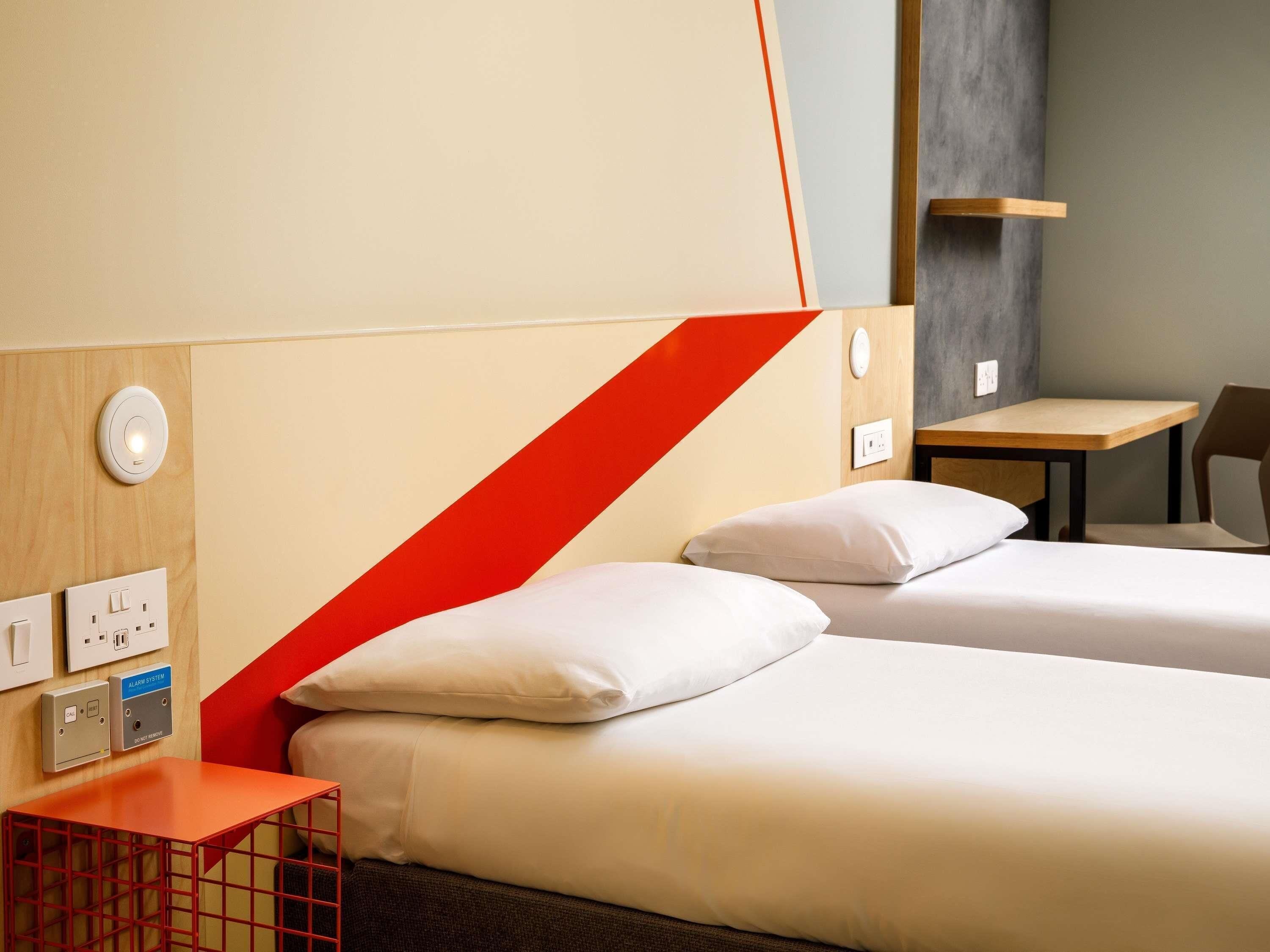 A hotel room with two beds, a small table, a chair, and a red accent wall.