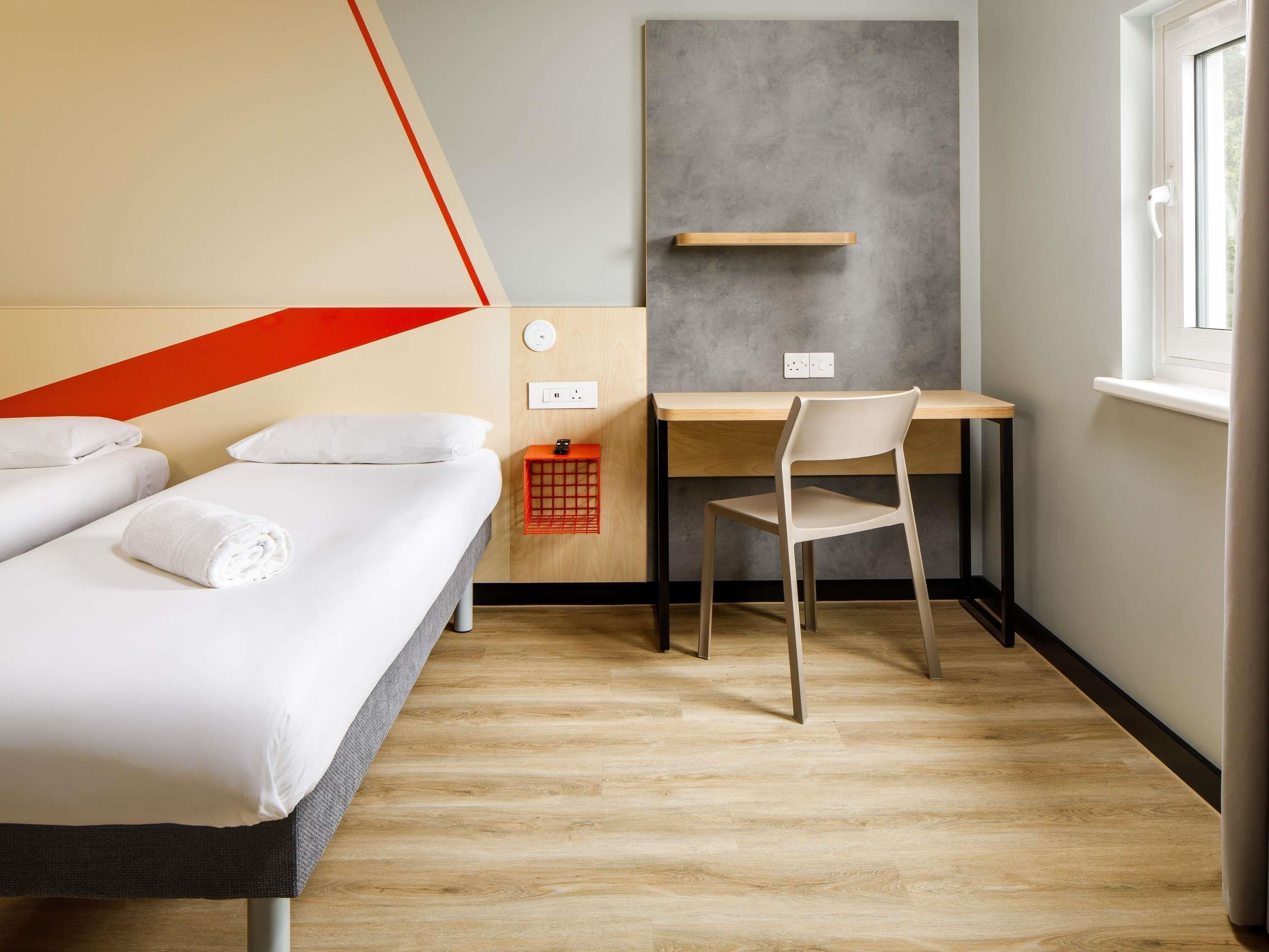 Habitación de hotel con dos camas, un escritorio, una silla, una cesta roja y una ventana.