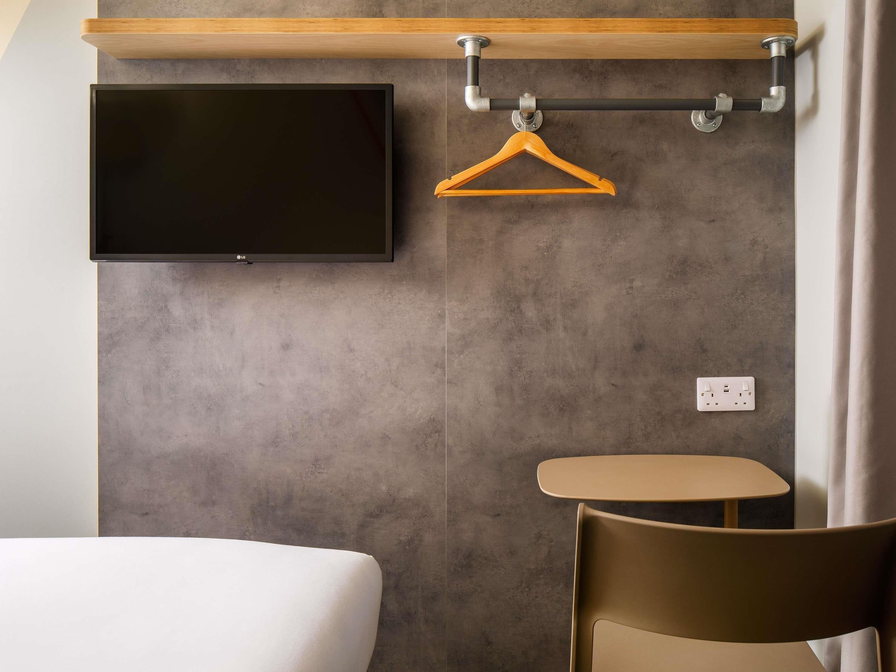 A modern hotel room with a flat-screen TV mounted on the wall, a wooden hanger, a small round table, and a single chair.