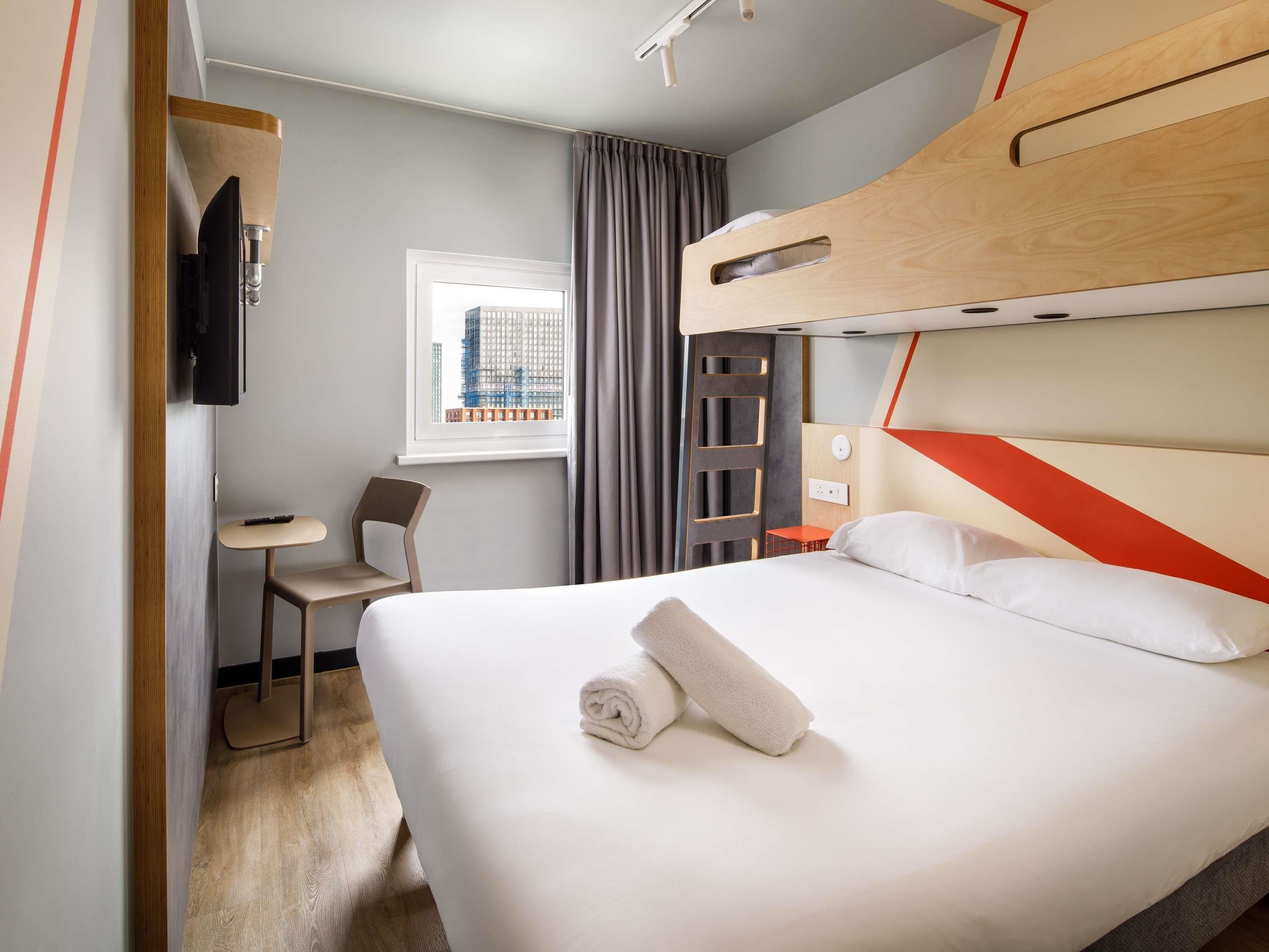 A hotel room with a bunk bed, a desk, a chair, a TV, and a window with a city view.