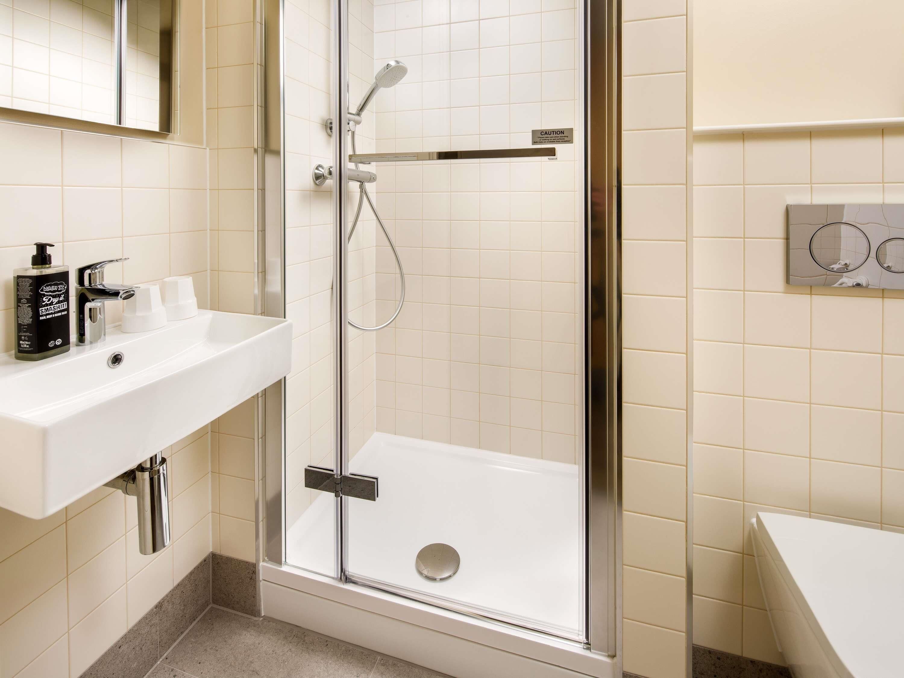 A modern bathroom with a glass-enclosed shower, a white sink with a mirror above it, and a wall-mounted control panel.