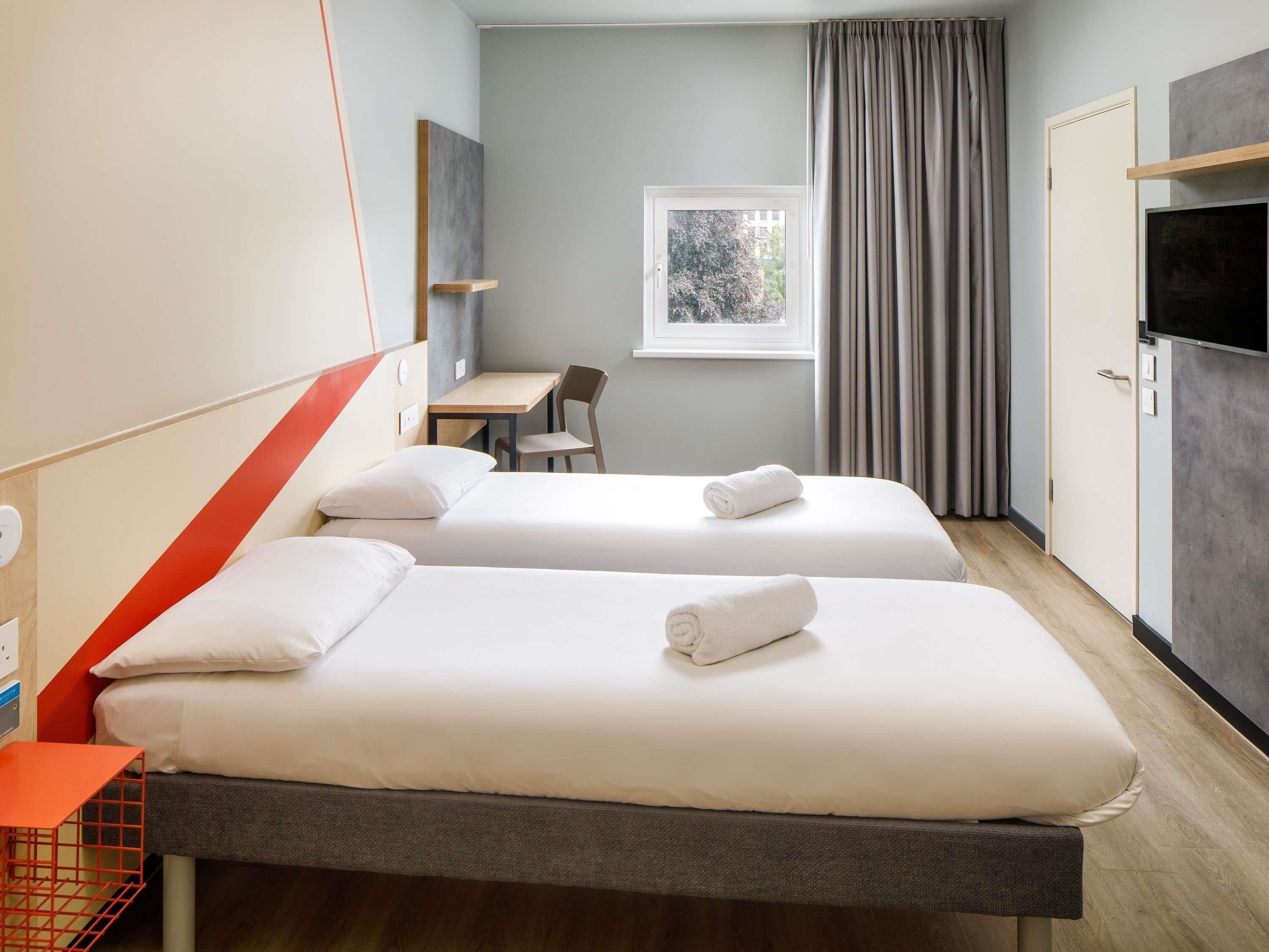 A hotel room with two beds, a desk, a chair, a television, and a window with curtains.