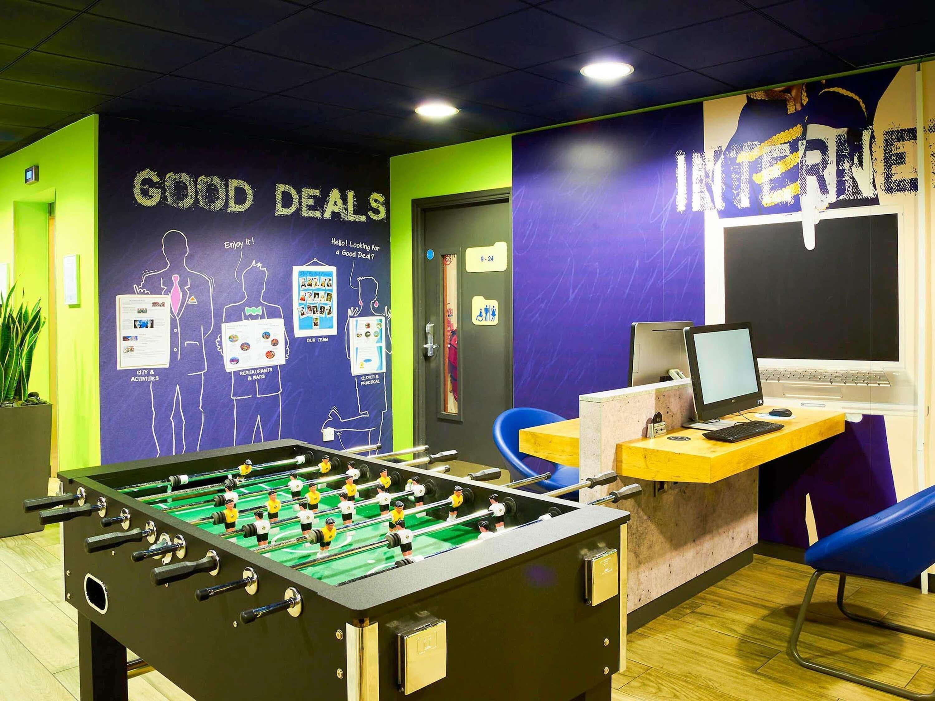 A room with a foosball table, a desk with a computer, and a wall with 'GOOD DEALS' and 'INTERNE' signs.
