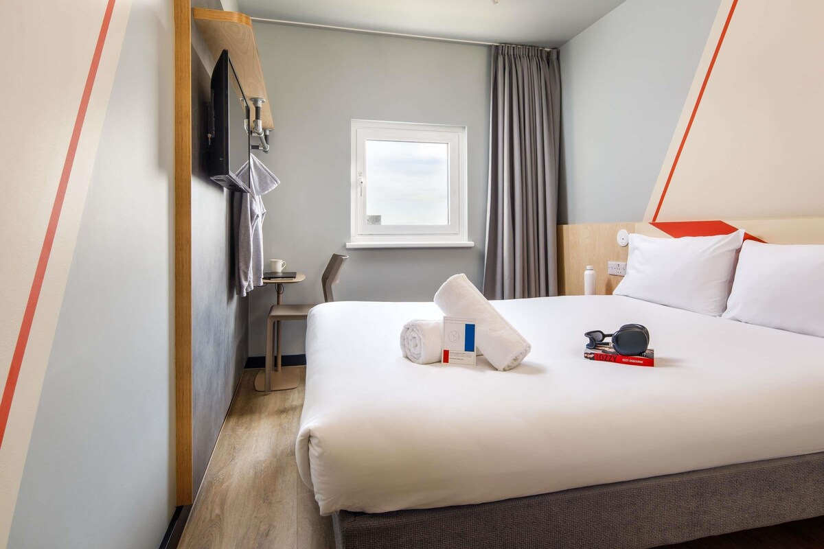 A hotel room with a bed, a desk, a chair, a window, and a TV mounted on the wall.